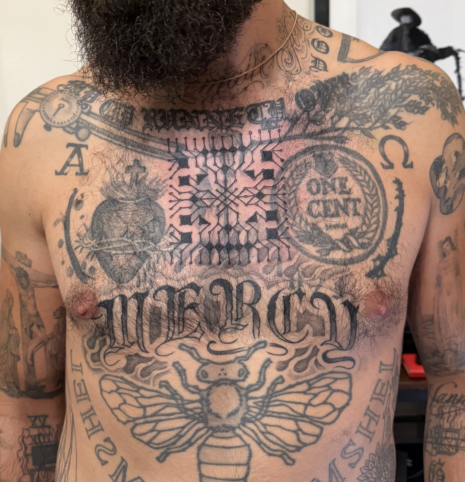 A large symmetrical geometric ornament tattooed in black across the centre of a client's chest, built from stepped blocks, fine radiating lines and dotted terminals, sitting between existing tattoos.