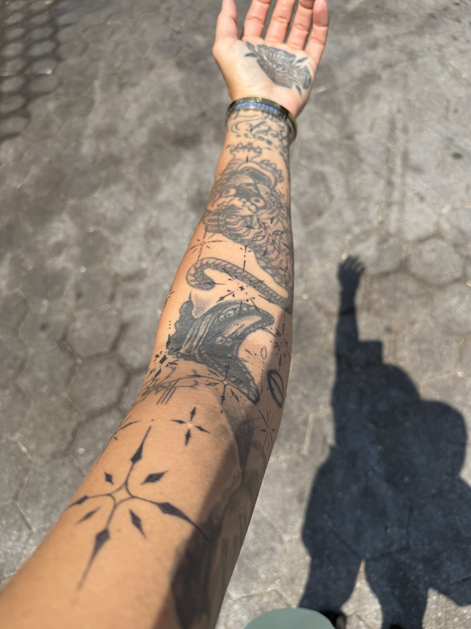 An outstretched forearm in sunlight showing ornamental blackwork: a large four-point motif with tapered diamond rays near the elbow, and smaller sparks and fine linework running down toward the wrist among existing tattoos.