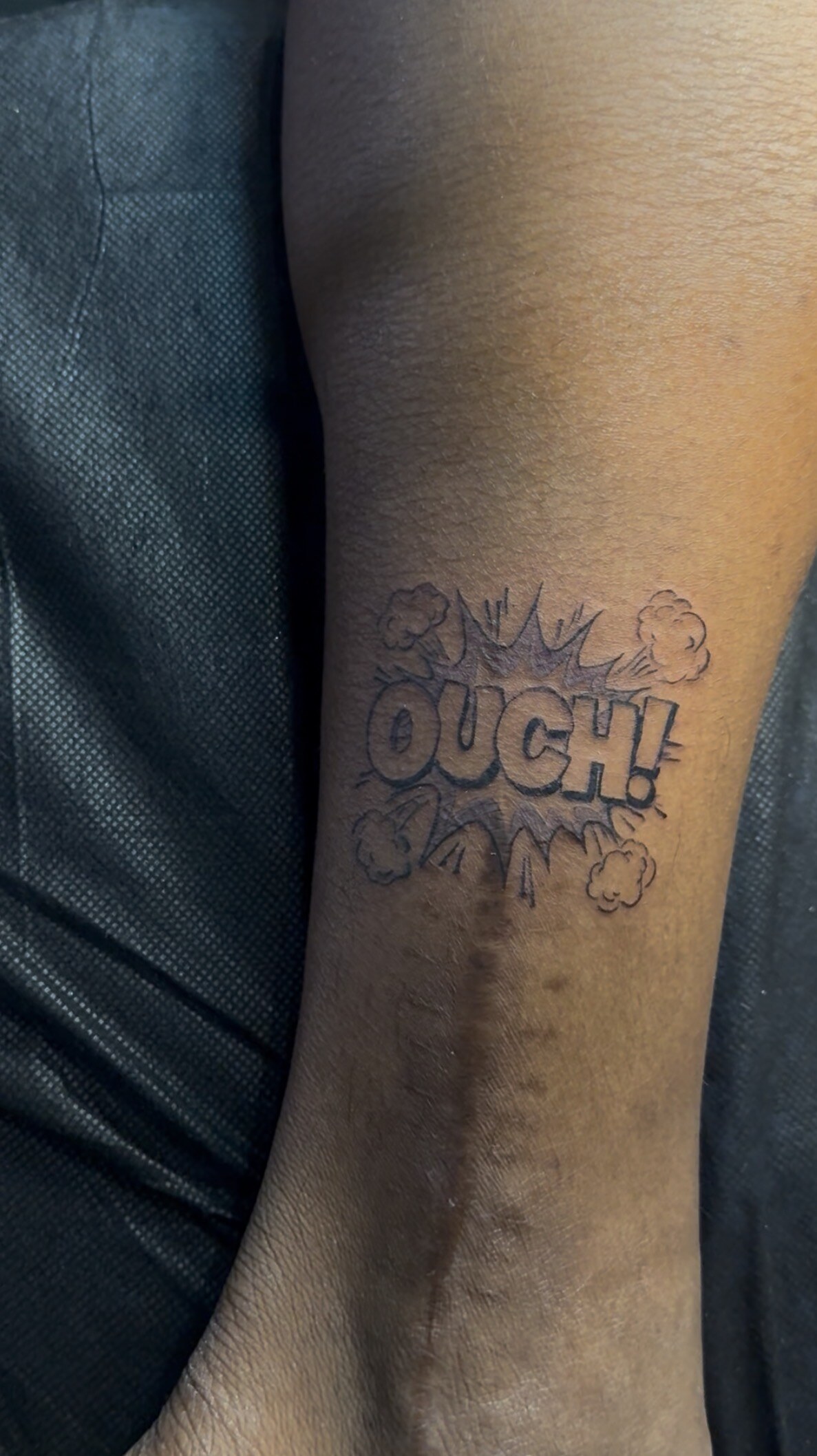 A comic-book style “OUCH!” burst tattooed in black and grey on the back of an upper arm.