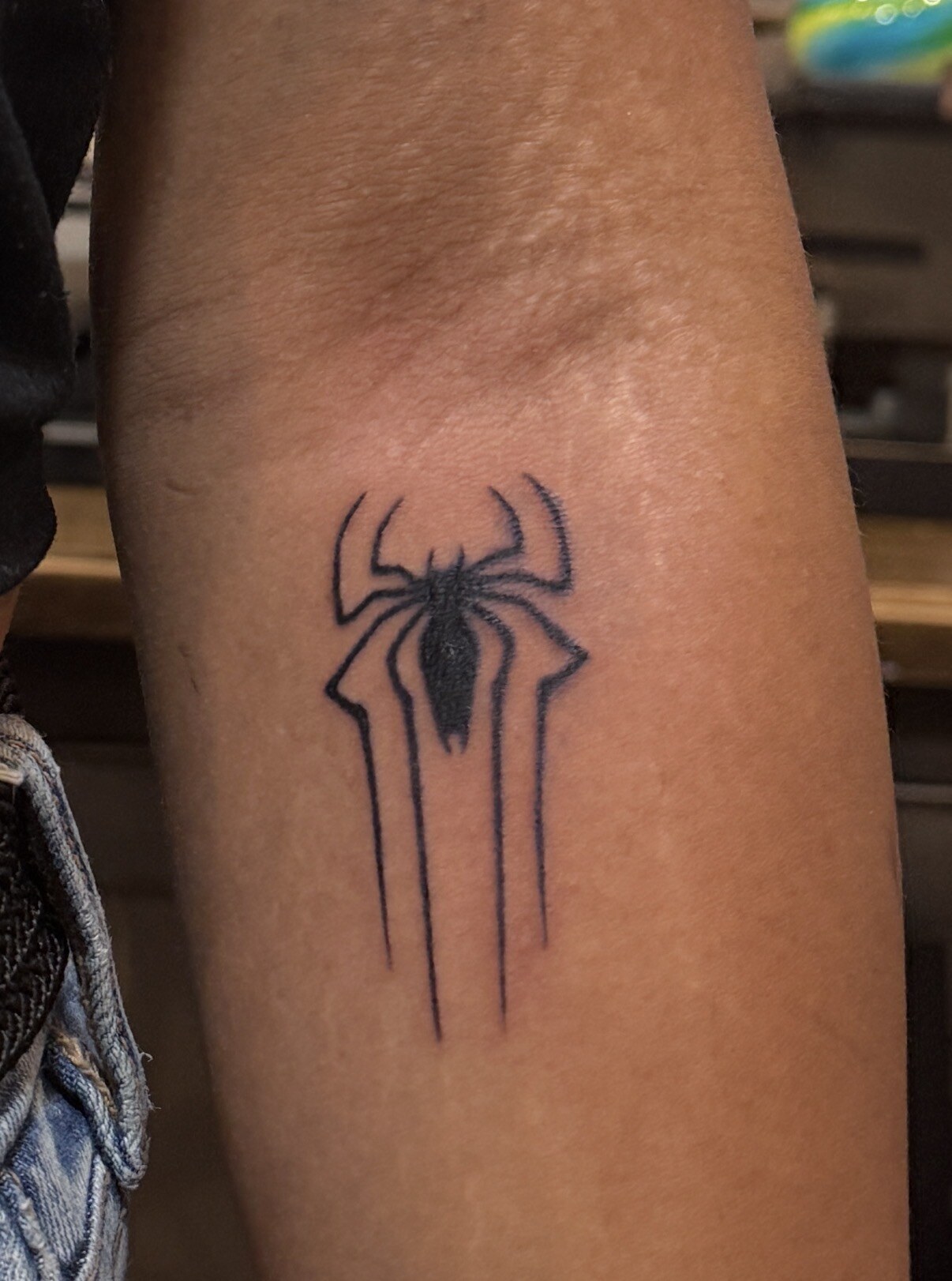A spider emblem tattooed in black on a forearm, with a solid black body, fine tapering legs, and long vertical lines trailing down beneath it.